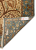 A close-up of a patterned area rug corner showing the underside with fringe, a blue-green field, and a cream border. A woven label is visible near the folded corner.