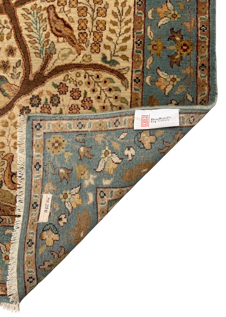 A close-up of a patterned area rug corner showing the underside with fringe, a blue-green field, and a cream border. A woven label is visible near the folded corner.