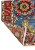 A close-up of a traditional patterned area rug with one corner folded over to show the underside and fringe. The rug features a blue field with red, green, and beige floral medallions and detailed borders.