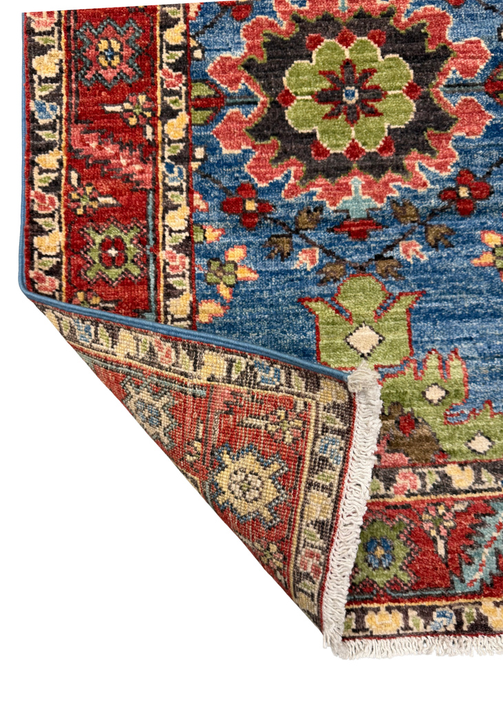 A close-up of a traditional patterned area rug with one corner folded over to show the underside and fringe. The rug features a blue field with red, green, and beige floral medallions and detailed borders.
