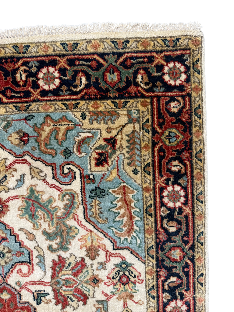 Close-up of an ornate patterned rug corner showing cream, navy, red, and light blue sections with woven floral motifs and a fringed edge.