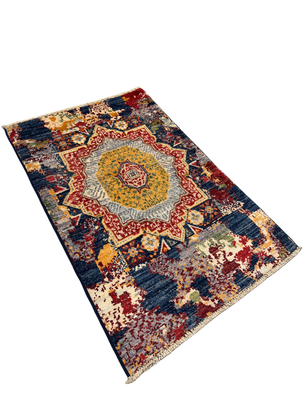 A rectangular area rug with a distressed, vintage-inspired pattern, featuring a large central medallion in yellow, red, and blue, surrounded by a patchwork of faded colors including navy, beige, and rust. The rug is shown at an angle on a plain white background.