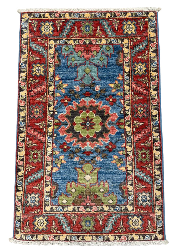 A rectangular hand-woven Persian-style area rug with a central blue field, red borders, and multicolored floral medallions, shown flat against a gray concrete floor.
