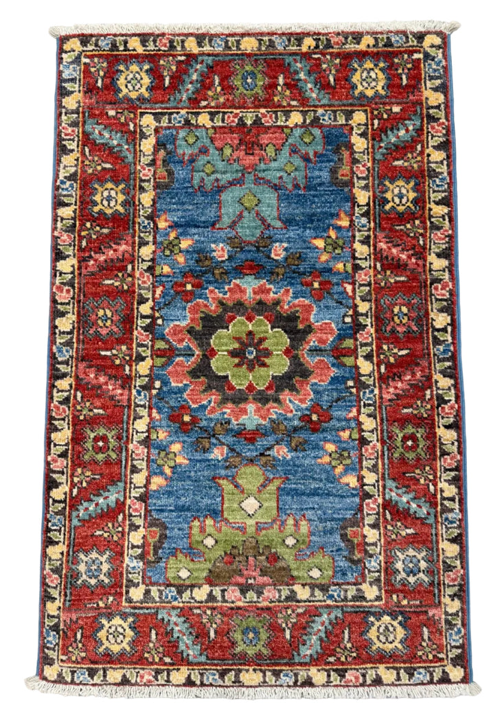 A rectangular hand-woven Persian-style area rug with a central blue field, red borders, and multicolored floral medallions, shown flat against a gray concrete floor.