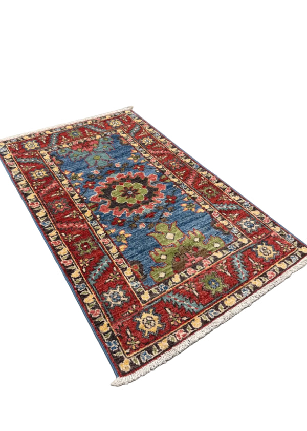 A small rectangular area rug with a traditional floral pattern, featuring a blue center, red borders, and cream fringe, displayed on a gray concrete floor.