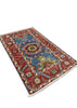 A small rectangular area rug with a traditional floral pattern, featuring a blue center, red borders, and cream fringe, displayed on a gray concrete floor.