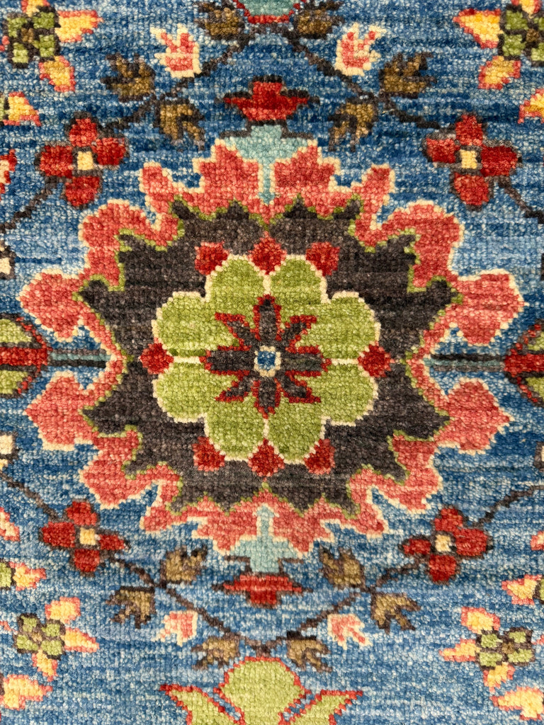 A close-up of a traditional patterned rug featuring a central medallion in light green and red, surrounded by coral and red floral motifs on a textured blue background.