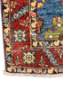 A close-up of a patterned area rug corner showing a red field with cream, blue, green, and black motifs, a blue section with a green cactus design, and white fringe along the bottom edge.