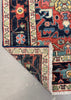 A close-up of a patterned area rug corner with one end folded back to show the underside and fringe, placed on a light gray floor.