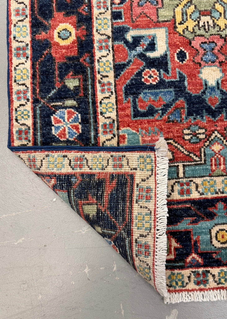 A close-up of a patterned area rug corner with one end folded back to show the underside and fringe, placed on a light gray floor.