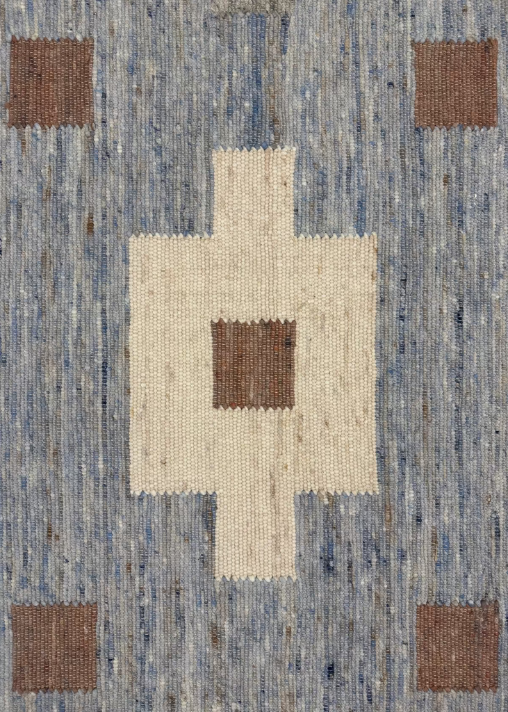 A close-up of a woven rug or textile with a geometric Swedish-style pattern. The design features a large cream-colored central diamond shape with a smaller brown square in the middle, surrounded by a textured blue-gray field with four matching brown corner squares.