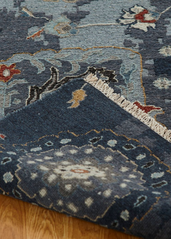 A close-up of a patterned area rug with a folded corner showing its cream fringe, set on a light brown wooden floor. The rug features a dark blue-gray base with traditional motifs in beige, light blue, and rust red.
