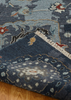 A close-up of a patterned area rug with a folded corner showing its cream fringe, set on a light brown wooden floor. The rug features a dark blue-gray base with traditional motifs in beige, light blue, and rust red.